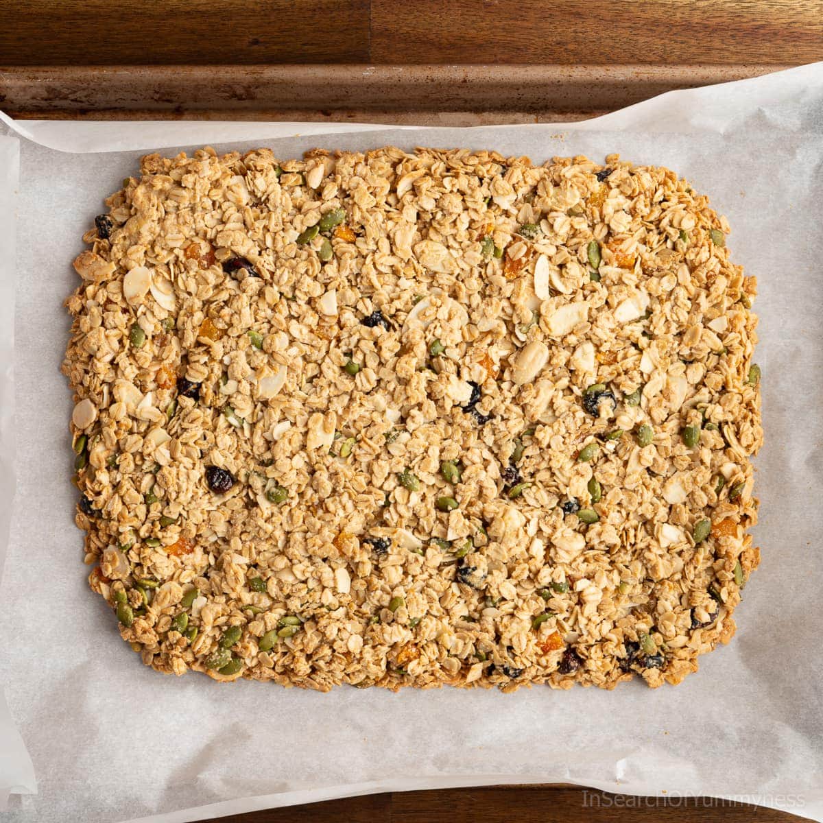 A golden-brown slab of baked homemade granola on a parchment-lined sheet pan that just came out of the oven.