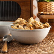 A grey bowl full of homemade granola on a woven placement, next to two spoons and another grey bowl.