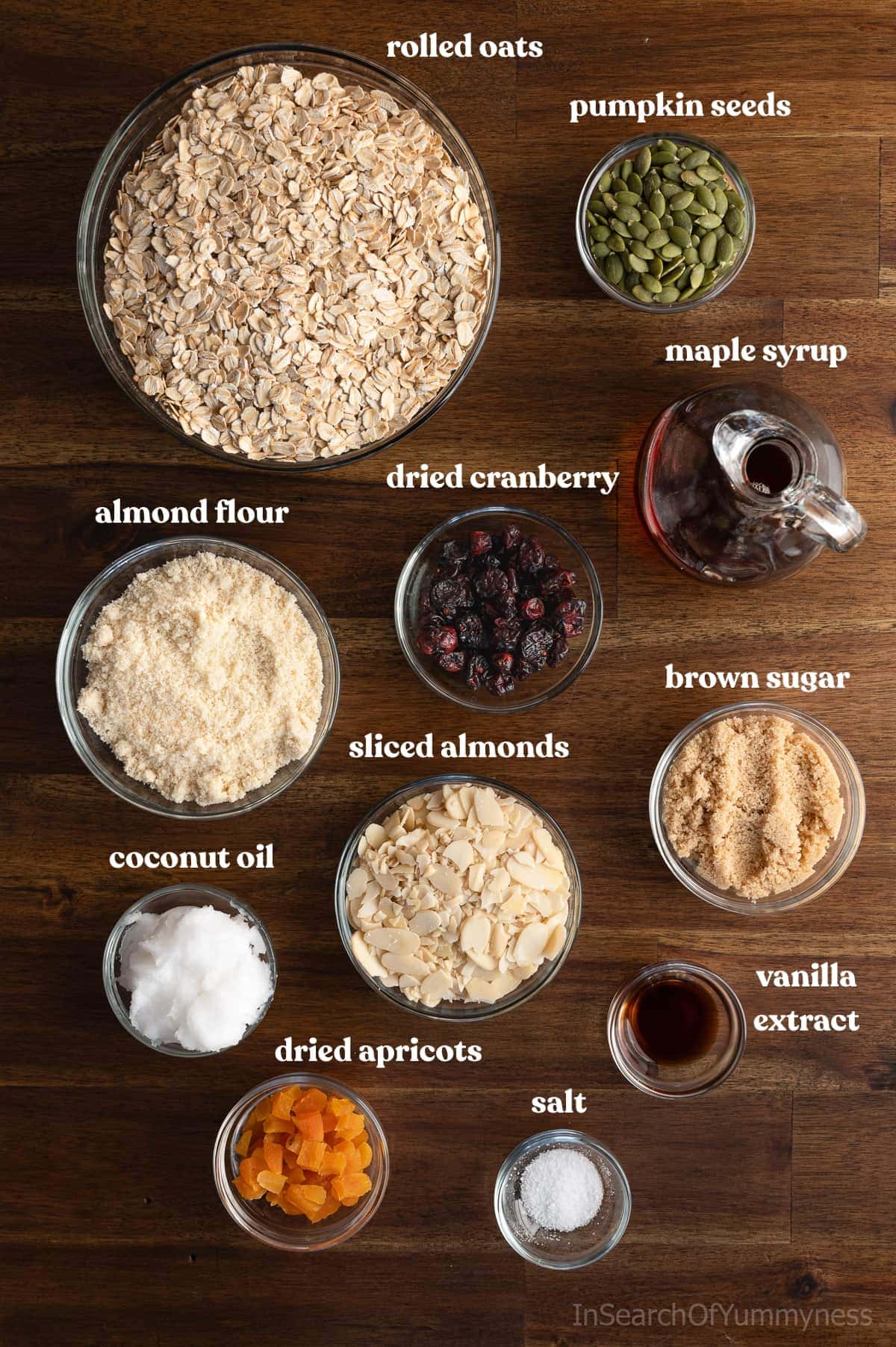 Ingredients for homemade granola in clear bowls on a wooden table, alongside a clear pitcher of maple syrup.