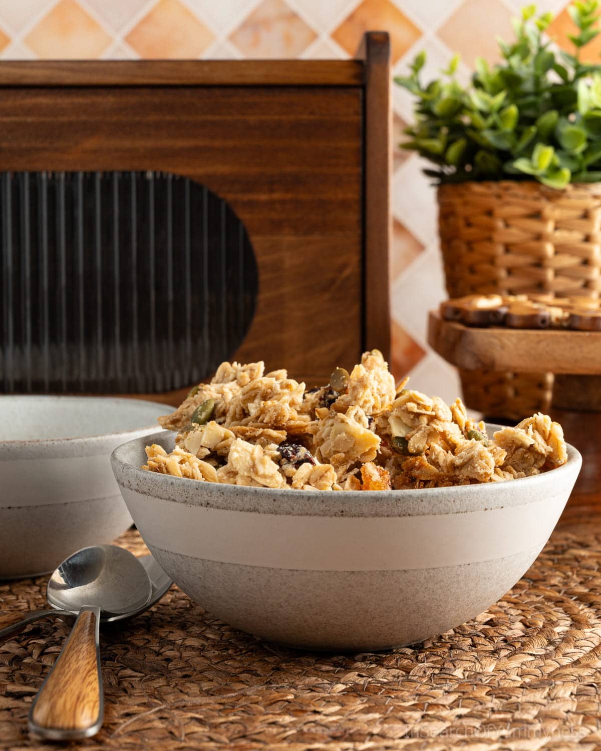 A grey bowl full of homemade granola on a woven placement, next to two spoons and another grey bowl.