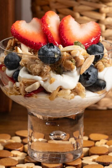 A wide dessert glass filled with granola parfait ingredients, including yogurt, honey, granola, almonds, fresh blueberries and fresh strawberries.