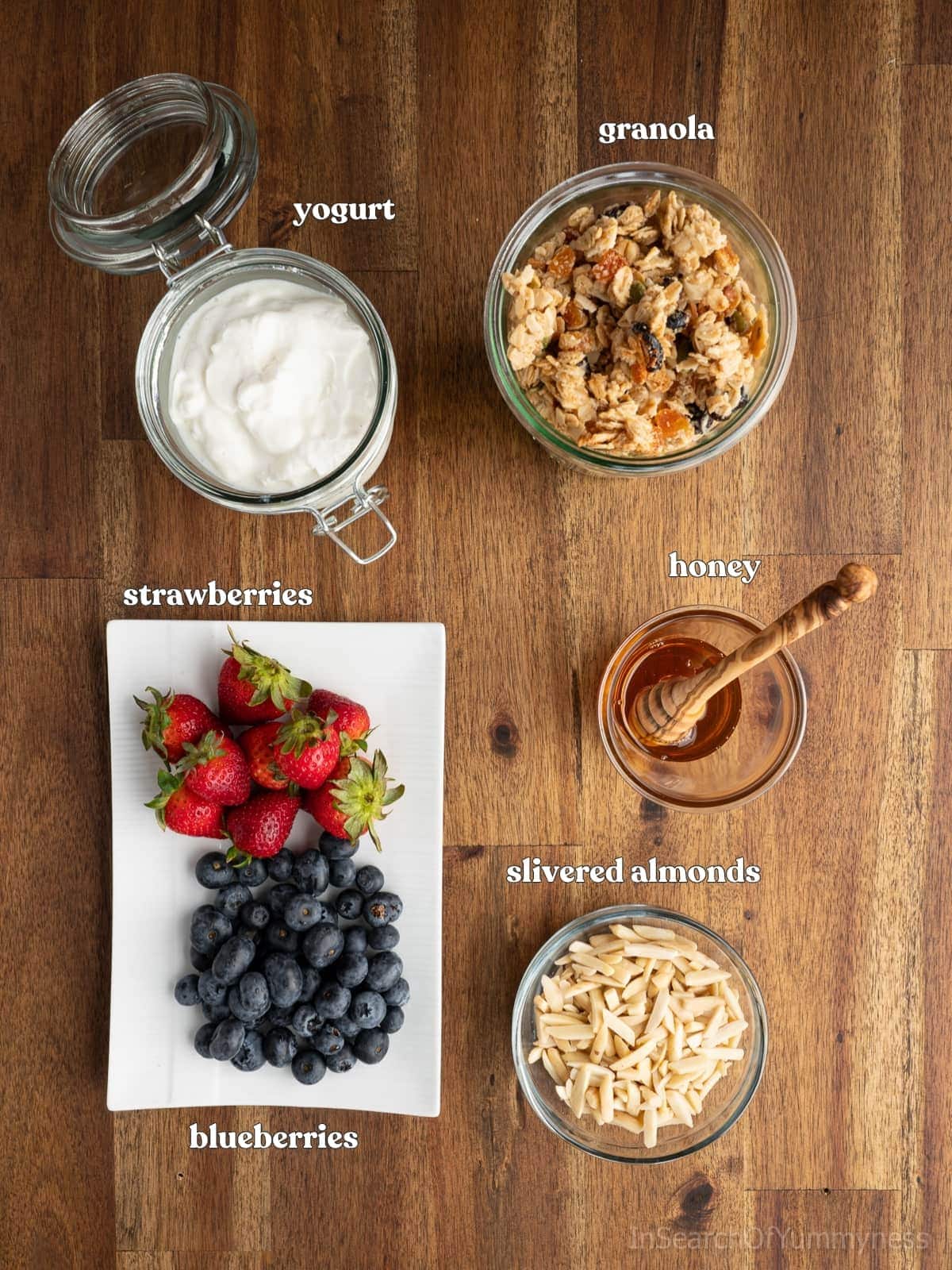 Yogurt in a glass jar, granola in a glass jar, honey in a glass with a honey dipper, a glass bowl of slivered almonds and a white platter with fresh blueberries and strawberries.