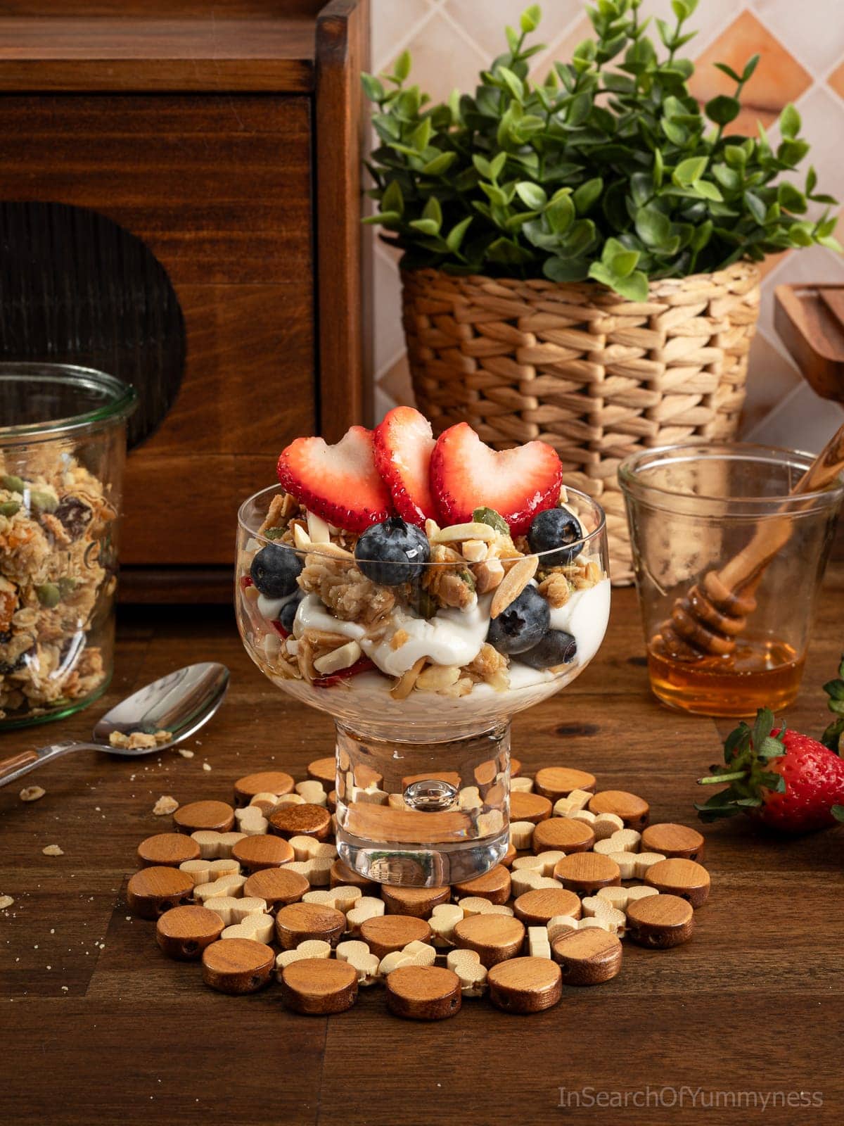 A granola parfait with berries in a wide dessert glass, on a table next to a jar of granola, a glass with honey and a honey dipper, and more strawberries on the table.