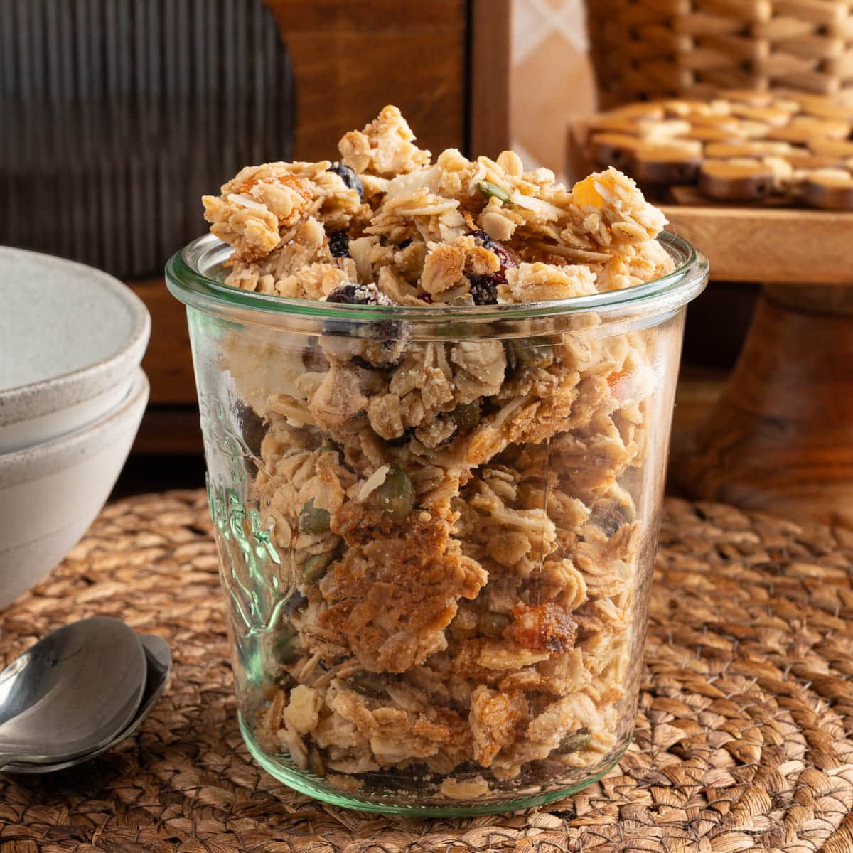 A weck jar full of simple homemade granola on a woven placemat next to two spoons and two stacked bowls.