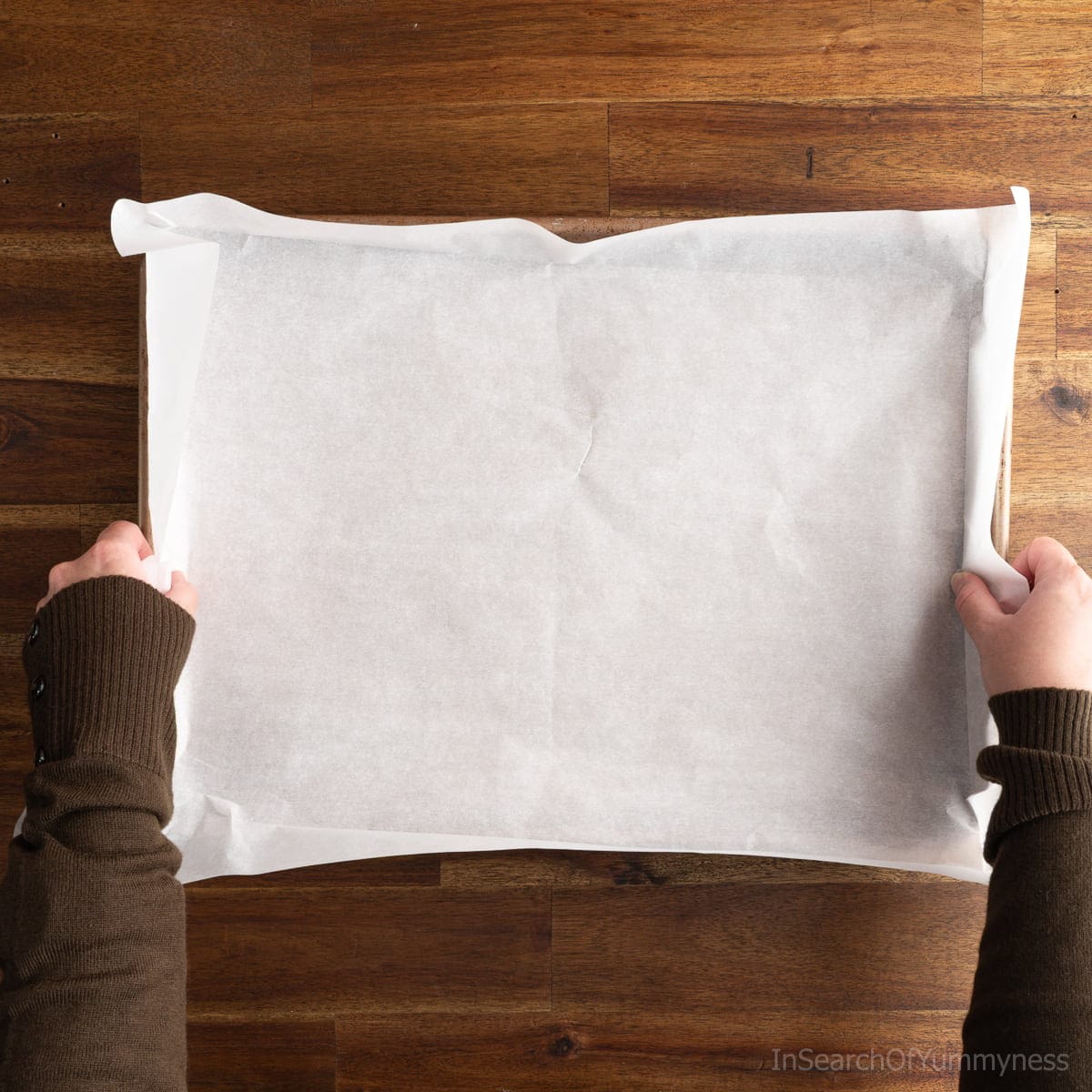 A pair of hands placing a piece of rectangular parchment paper over a metal sheet pan.