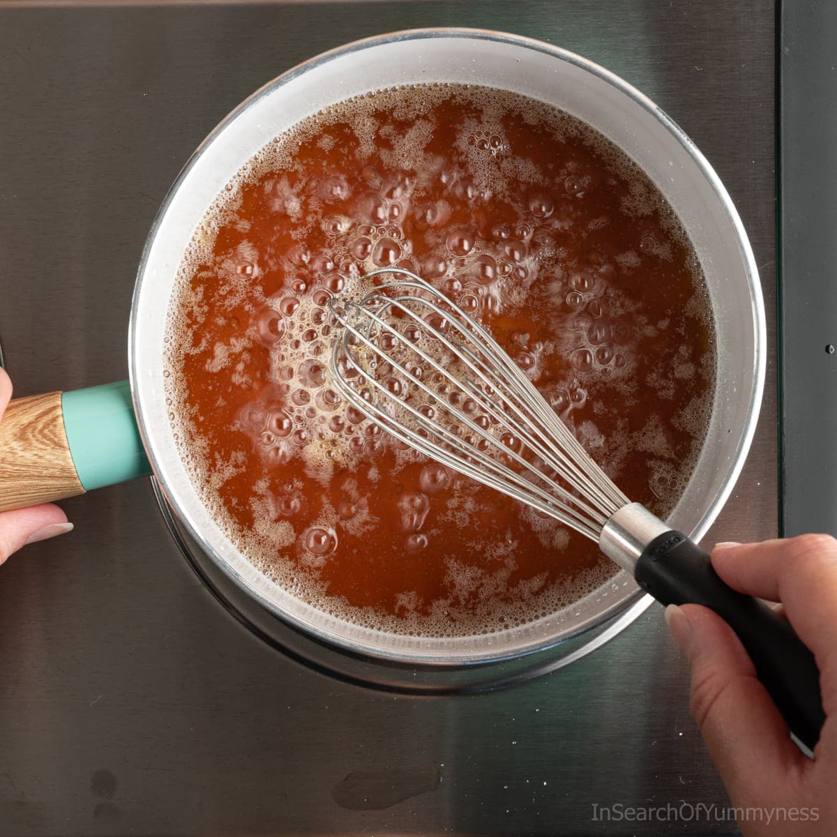The wet ingredients for homemade granola, including coconut oil and maple syrup, heating up in a small white pot. Someone is using a whisk to stir the mixture.