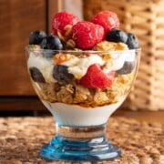 A maple parfait in a small dessert glass with a blue bottom. The parfait contains layers of yogurt, maple granola, maple syrup, sliced almonds and fresh berries.