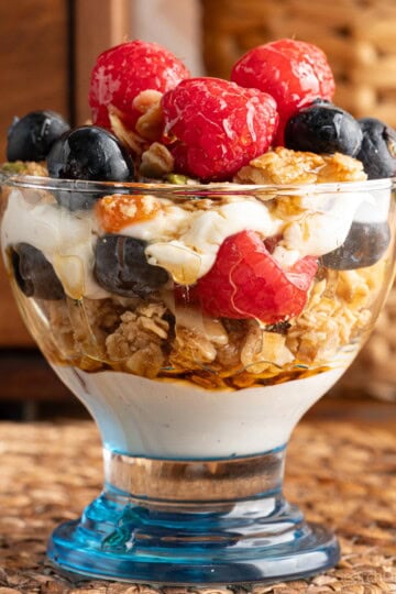 A maple parfait in a small dessert glass with a blue bottom. The parfait contains layers of yogurt, maple granola, maple syrup, sliced almonds and fresh berries.