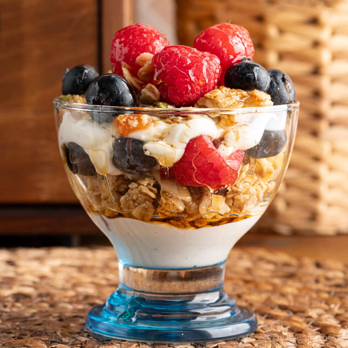 A maple parfait in a small dessert glass with a blue bottom. The parfait contains layers of yogurt, maple granola, maple syrup, sliced almonds and fresh berries.