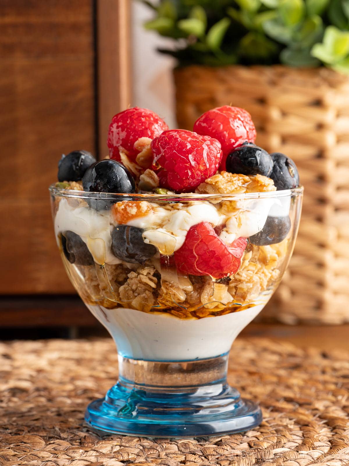 A maple parfait in a small dessert glass with a blue bottom. The parfait contains layers of yogurt, maple granola, maple syrup, sliced almonds and fresh berries.
