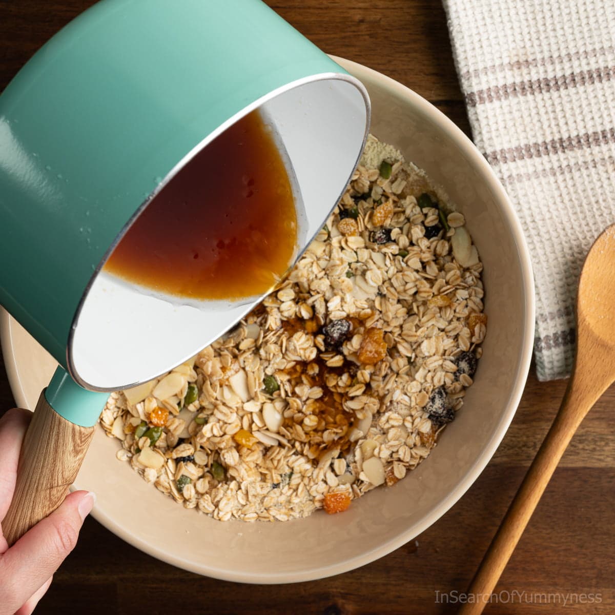 A hand pouring a maple syrup coconut oil syrup out of a small pot into a large bowl full of dry granola ingredients.