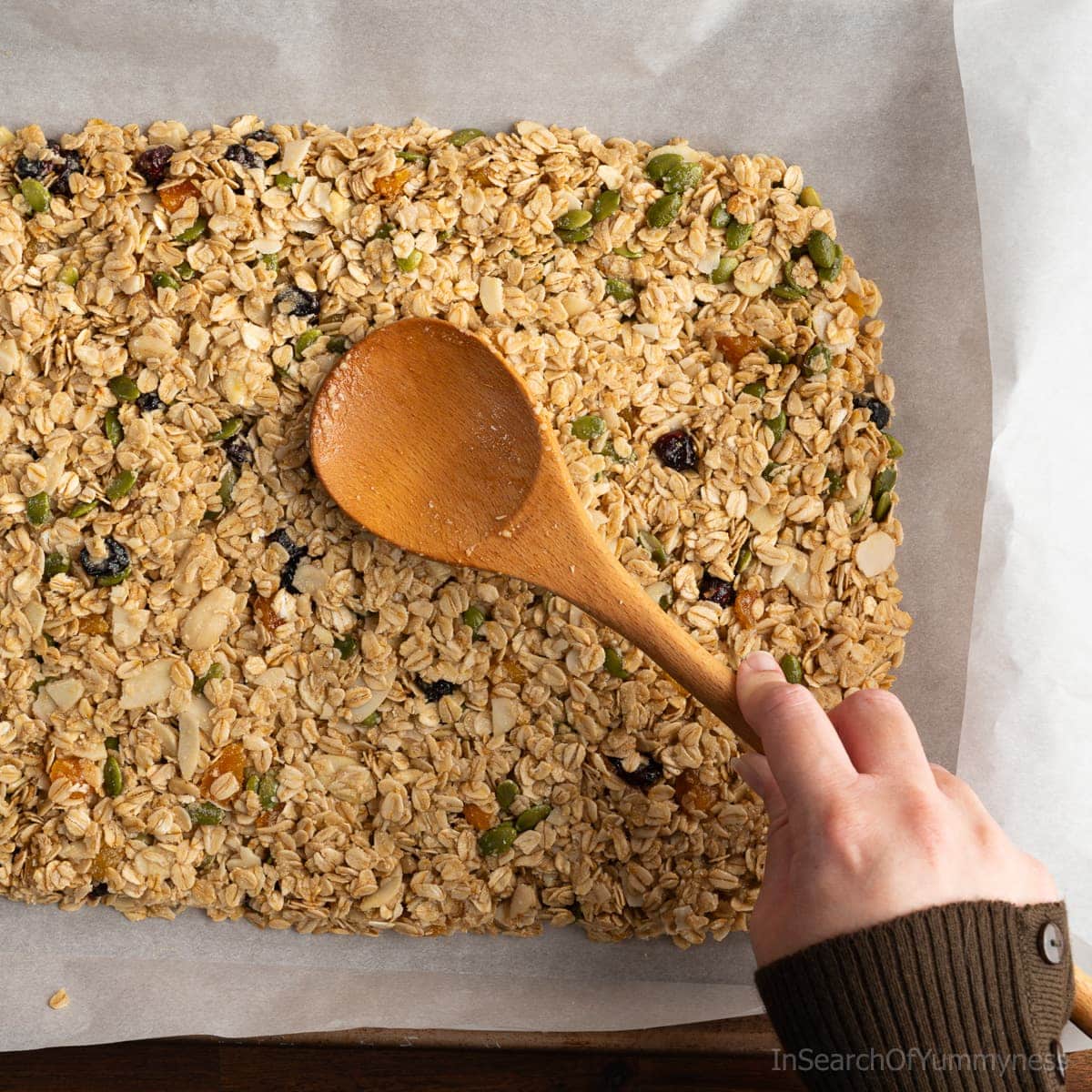 A hand using a wooden spoon to press a granola mixture into an even layer on a parchment-lined baking tray.