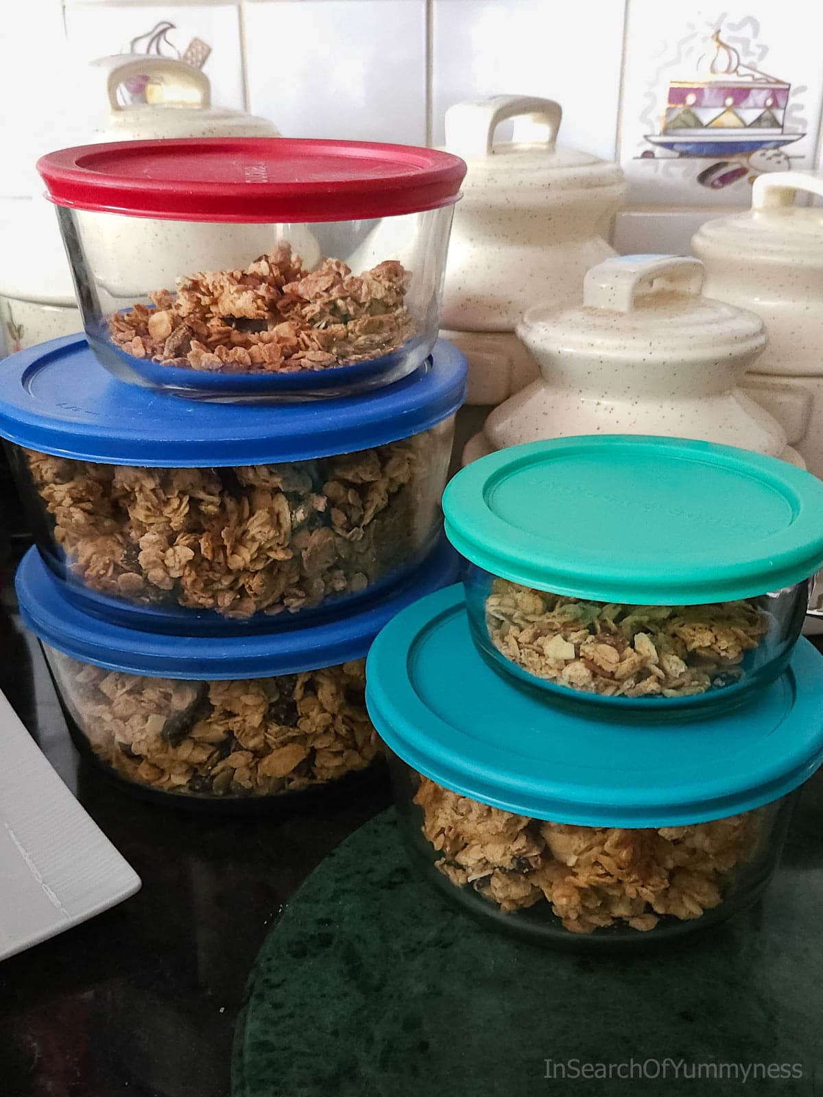 5 glass containers with colourful lids full of various versions of homemade granola on a dark countertop in a kitchen.