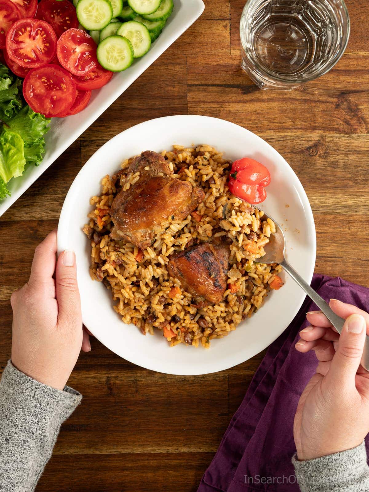 Someone taking a spoonful of Trinidadian pelau with chicken from a white plate. There is a salad next to the plate, as well as a glass of water.
