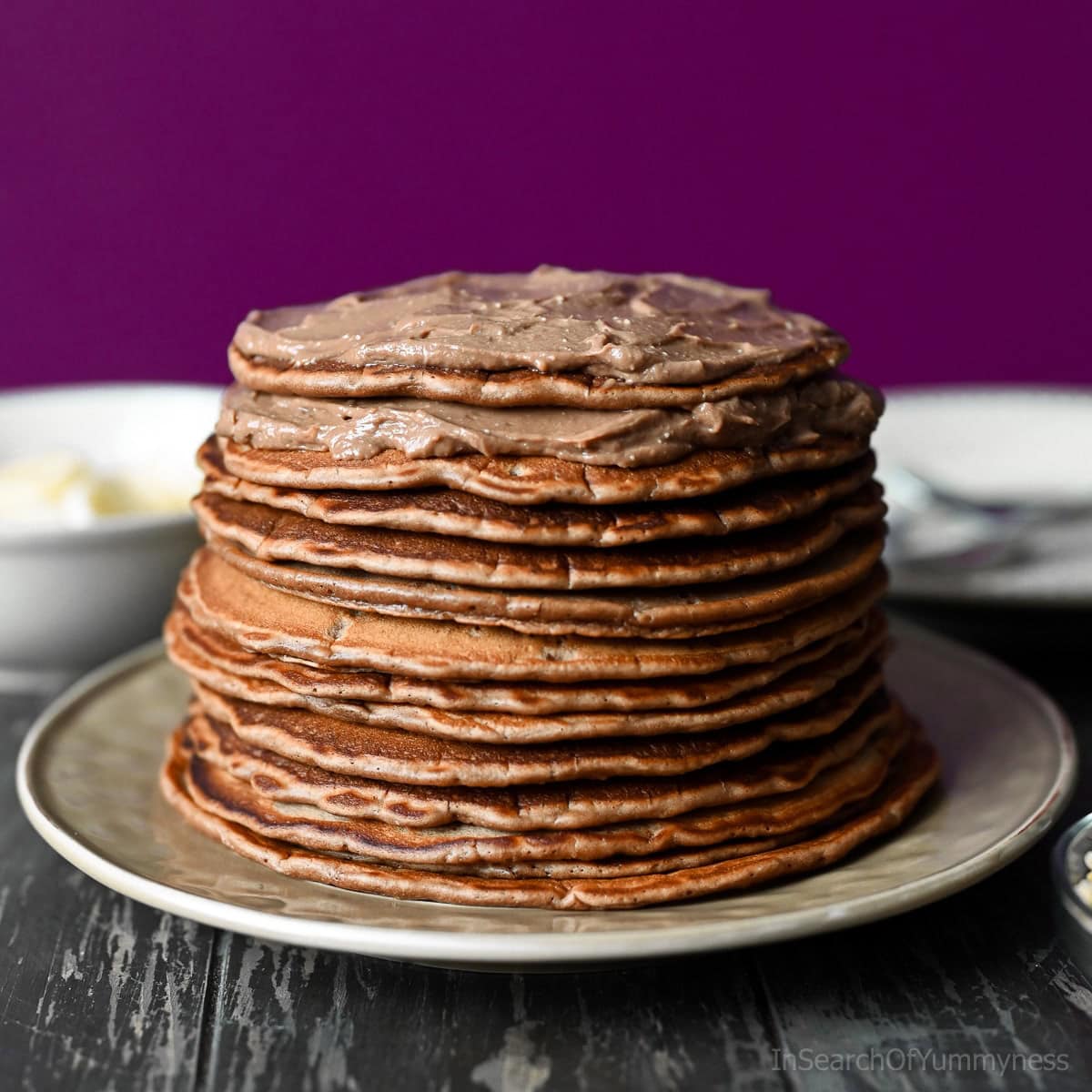 A large stack of Nutella pancakes on a beige plate on a grey wooden table.
