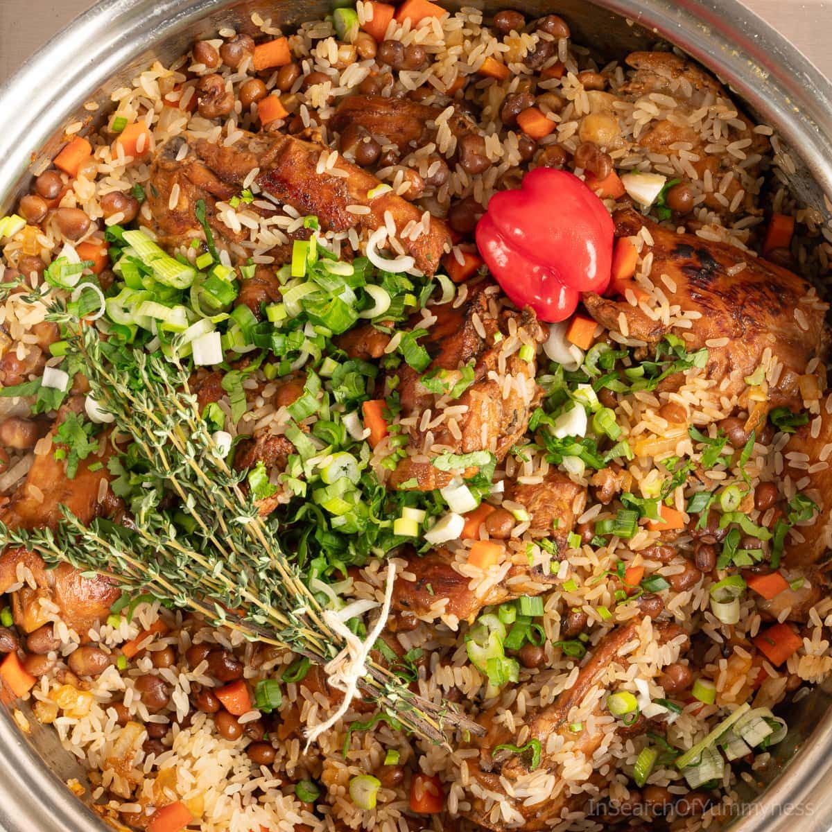 A stainless steel pot full of browned chicken, rice, carrots and onions, topped with chopped green onions and a sprig of thyme.
