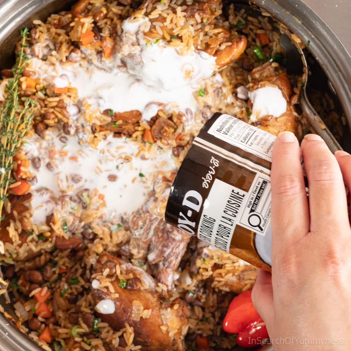 Adding canned coconut milk to a stainless steel pot with ingredients to make Trinidadian chicken pelau.