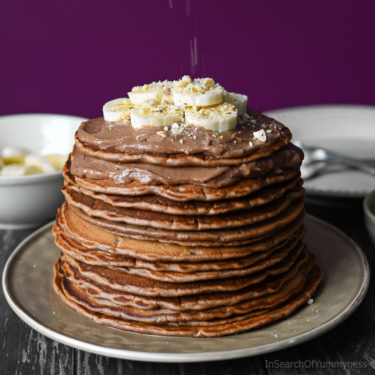 A large stack of cooked Nutella pancakes topped with mascarpone cream, sliced bananas and chopped hazelnuts.