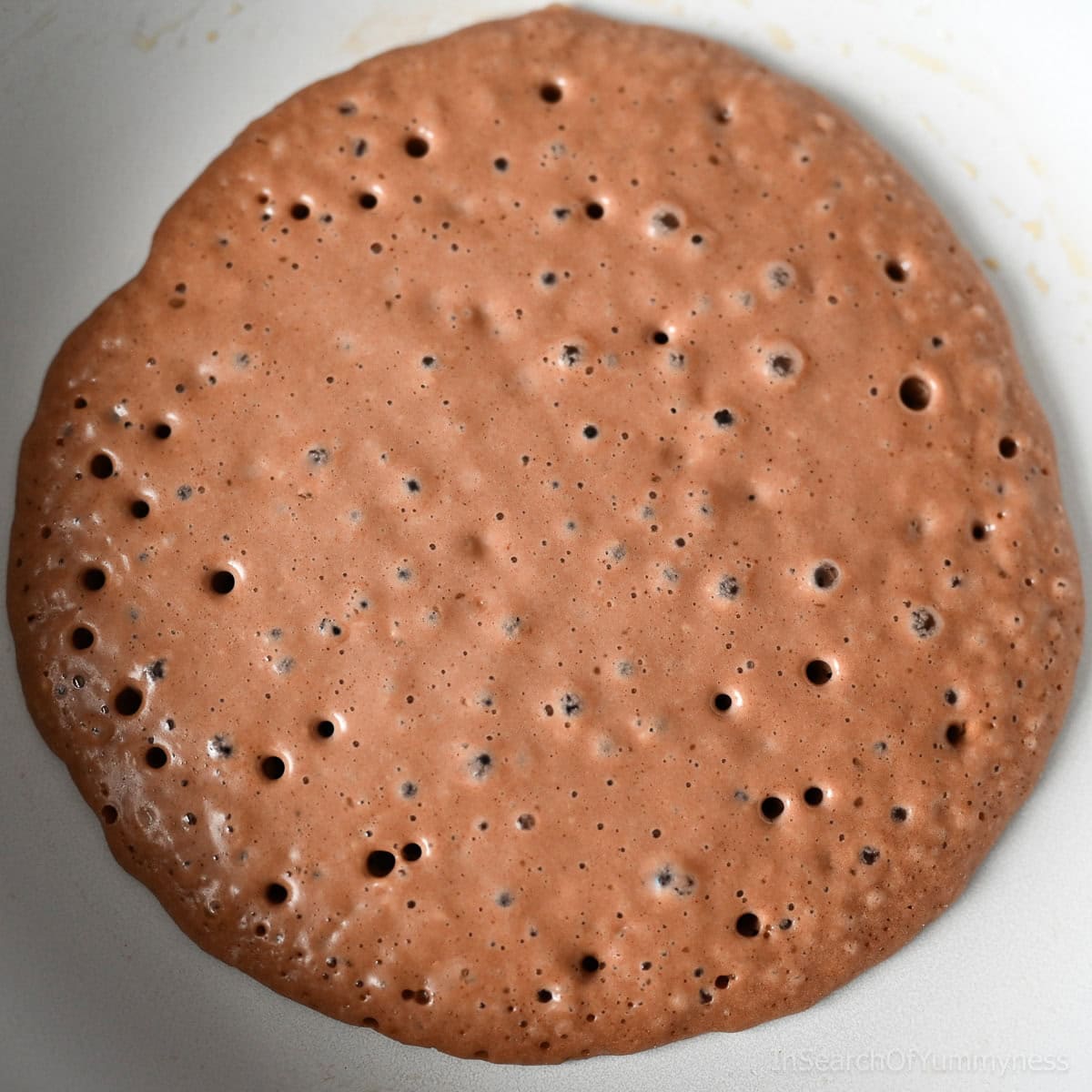 A Nutella pancake cooking in a white non-stick frying pan. Bubbles on top of the pancake indicate that it's time to flip it.