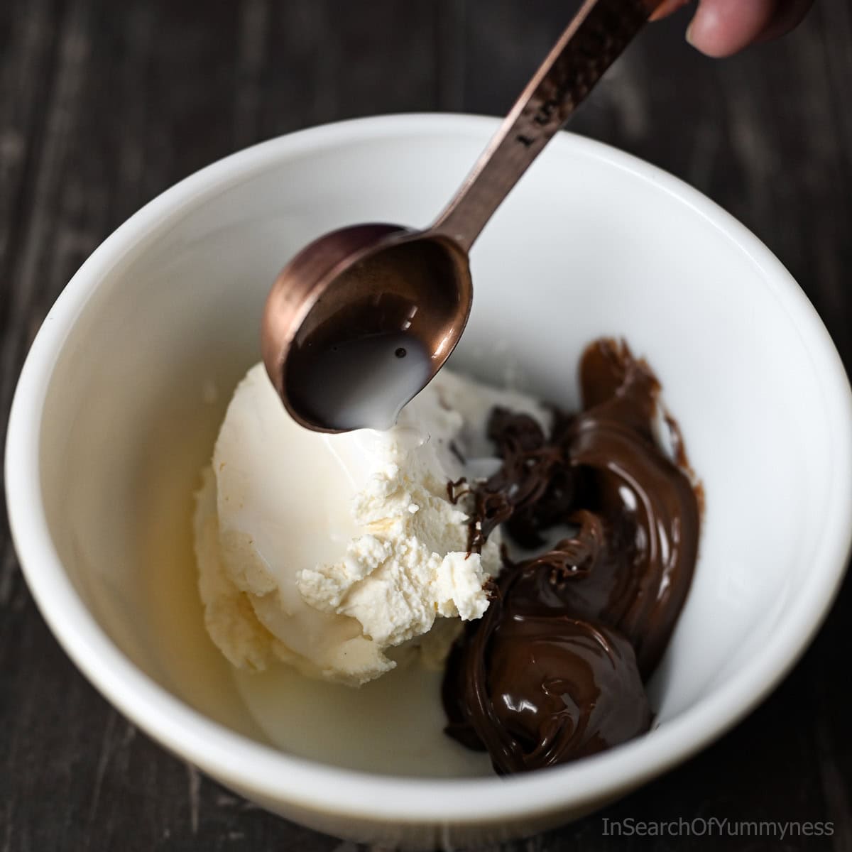 Milk being added from a measuring spoon into a white mixing bowl that already has mascarpone cheese and Nutella in it.