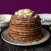 A tall stack of Nutella pancakes topped with mascarpone cream, sliced bananas and finely chopped hazelnuts on a beige plate.