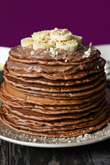 A tall stack of Nutella pancakes topped with mascarpone cream, sliced bananas and finely chopped hazelnuts on a beige plate.