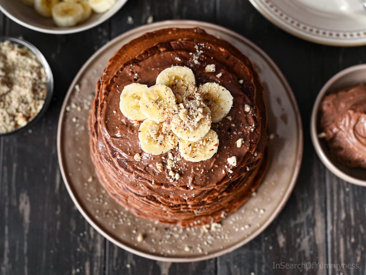 Looking down at a stack of Nutella pancakes topped with slices of fresh banana and finely diced hazelnuts, sitting on a beige plate on a dark grey table.
