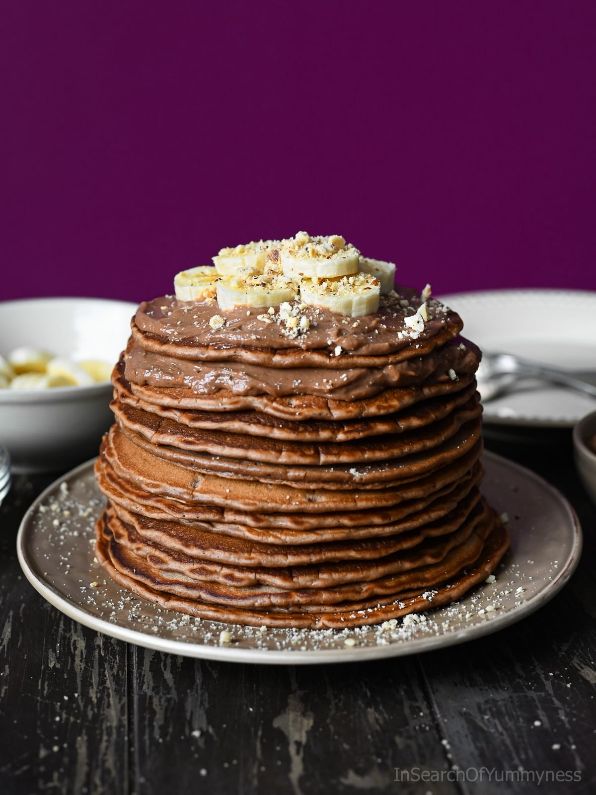A tall stack of Nutella pancakes topped with mascarpone cream, sliced bananas and finely chopped hazelnuts on a beige plate.