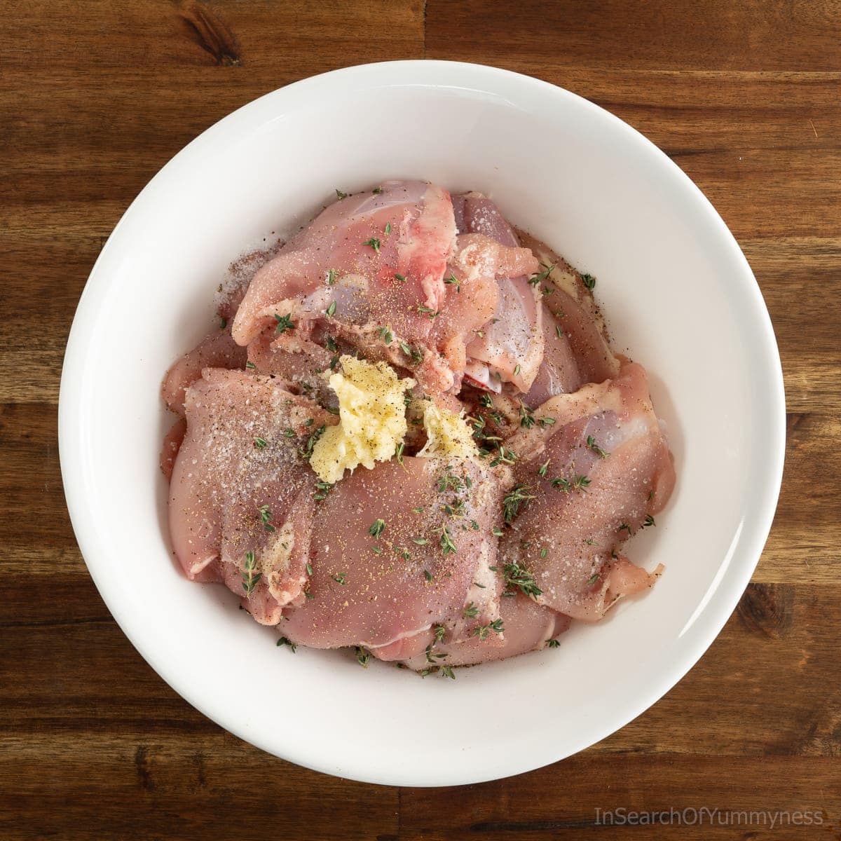 A white bowl full of raw skinless chicken thighs, sprinkled with fresh thyme, salt, pepper and fresh garlic.