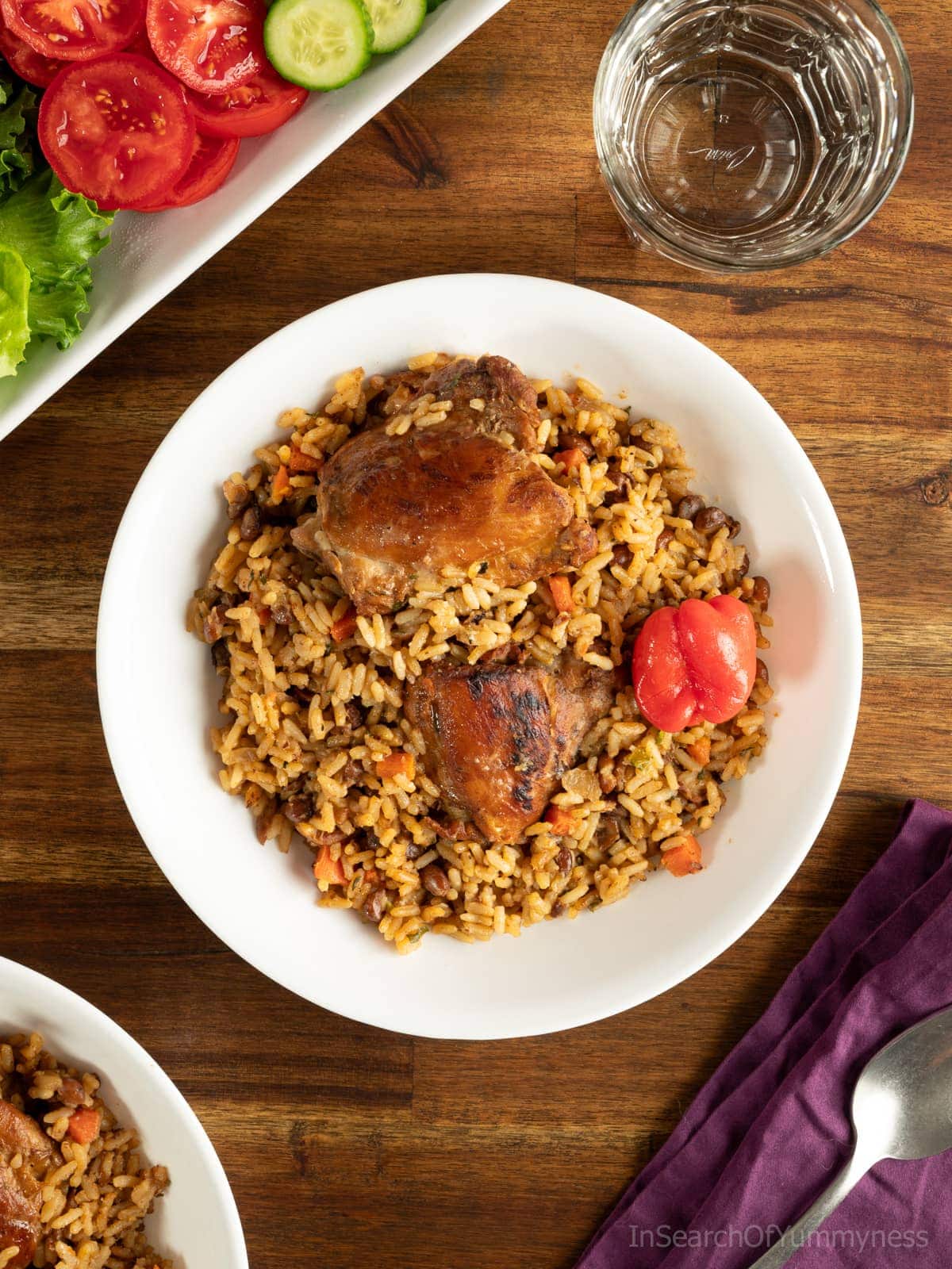 Trinidad pelau with chicken on a white plate, next to a purple napkin with a spoon resting on it. There is a salad next to the plate, as well as a glass of water.