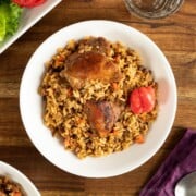 Trinidad pelau with chicken on a white plate, next to a purple napkin with a spoon resting on it. There is a salad next to the plate, as well as a glass of water.