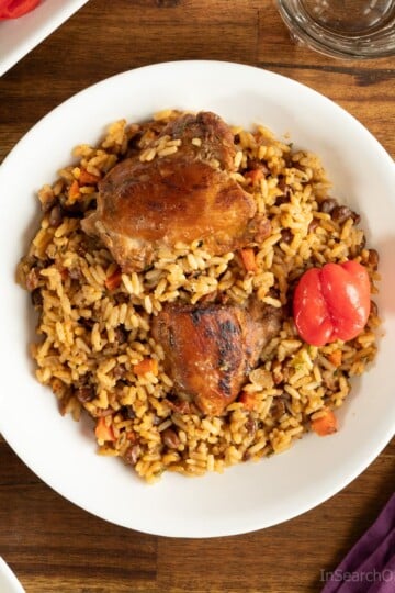 Trinidad pelau with chicken on a white plate, next to a purple napkin with a spoon resting on it. There is a salad next to the plate, as well as a glass of water.