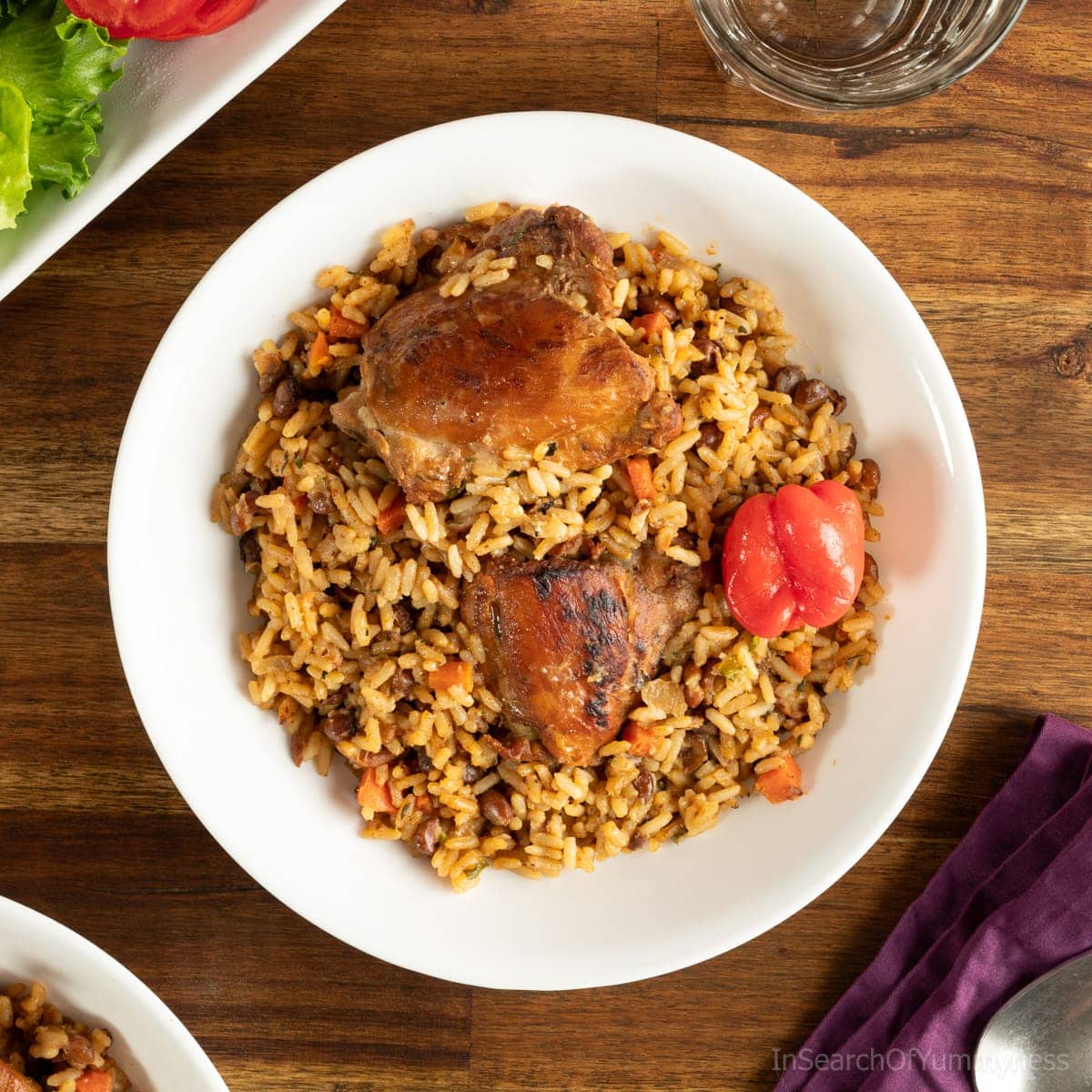 Trinidad pelau with chicken on a white plate, next to a purple napkin with a spoon resting on it. There is a salad next to the plate, as well as a glass of water.