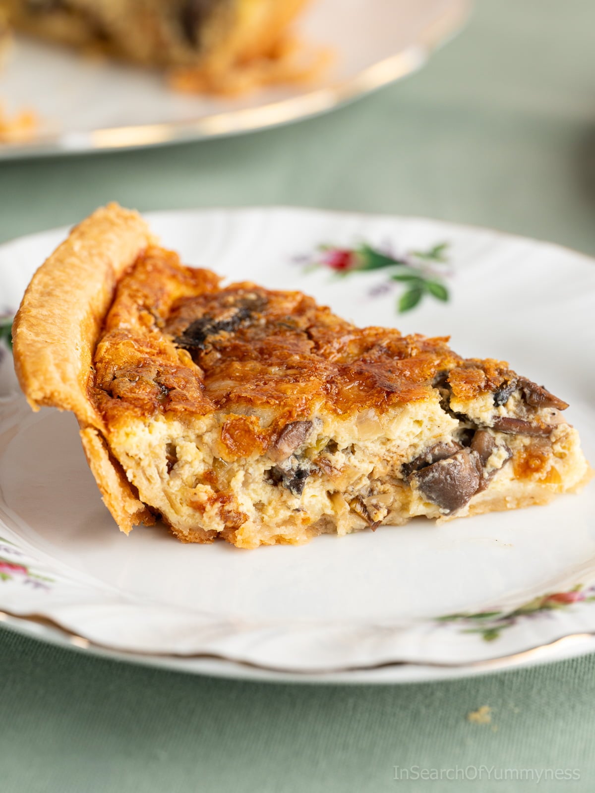 A slice of freshly baked leek and mushroom quiche with a cheddar cheese topping, sitting on a fancy white plate on a green tablecloth.