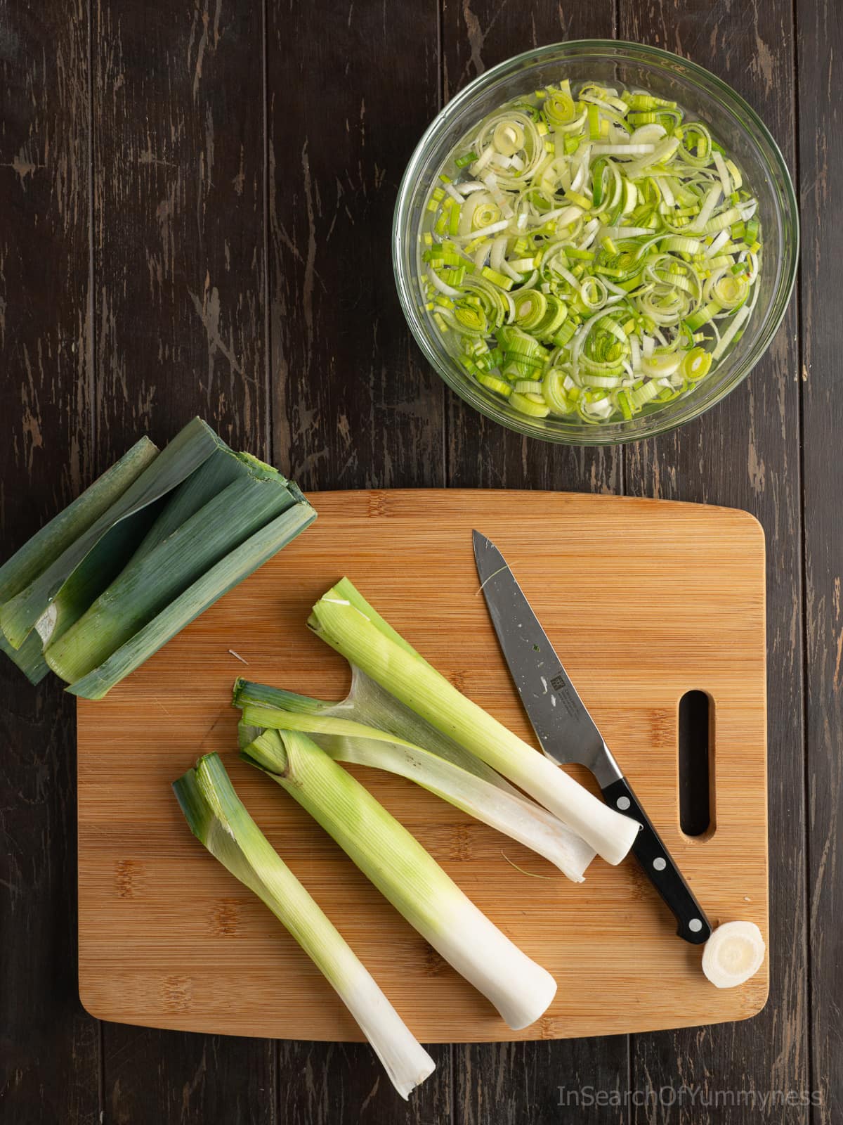 A leek cut into large pieces on a wooden cutting board next to a knife. You can see the dirt inside the leeks. There is a clear bowl of finely chopped leeks soaking in cold water above.
