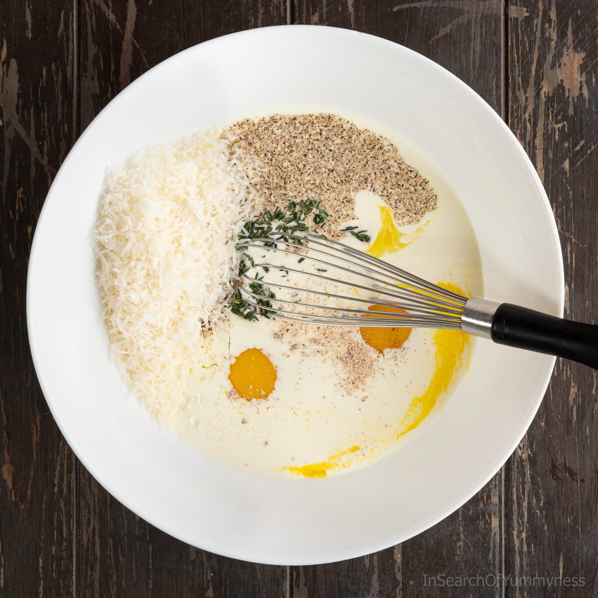 Eggs, milk, cream, seasonings and cheese in a large white bowl with a small whisk.