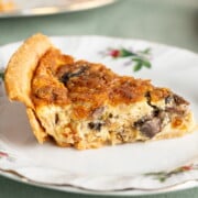 A slice of freshly baked leek and mushroom quiche with a cheddar cheese topping, sitting on a fancy white plate on a green tablecloth.