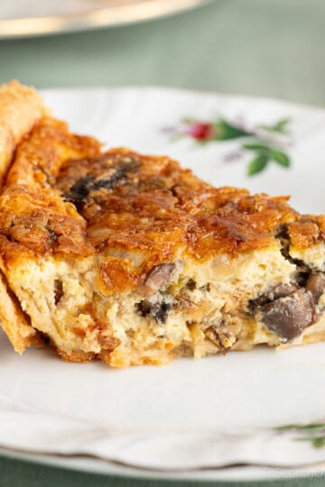 A slice of freshly baked leek and mushroom quiche with a cheddar cheese topping, sitting on a fancy white plate on a green tablecloth.