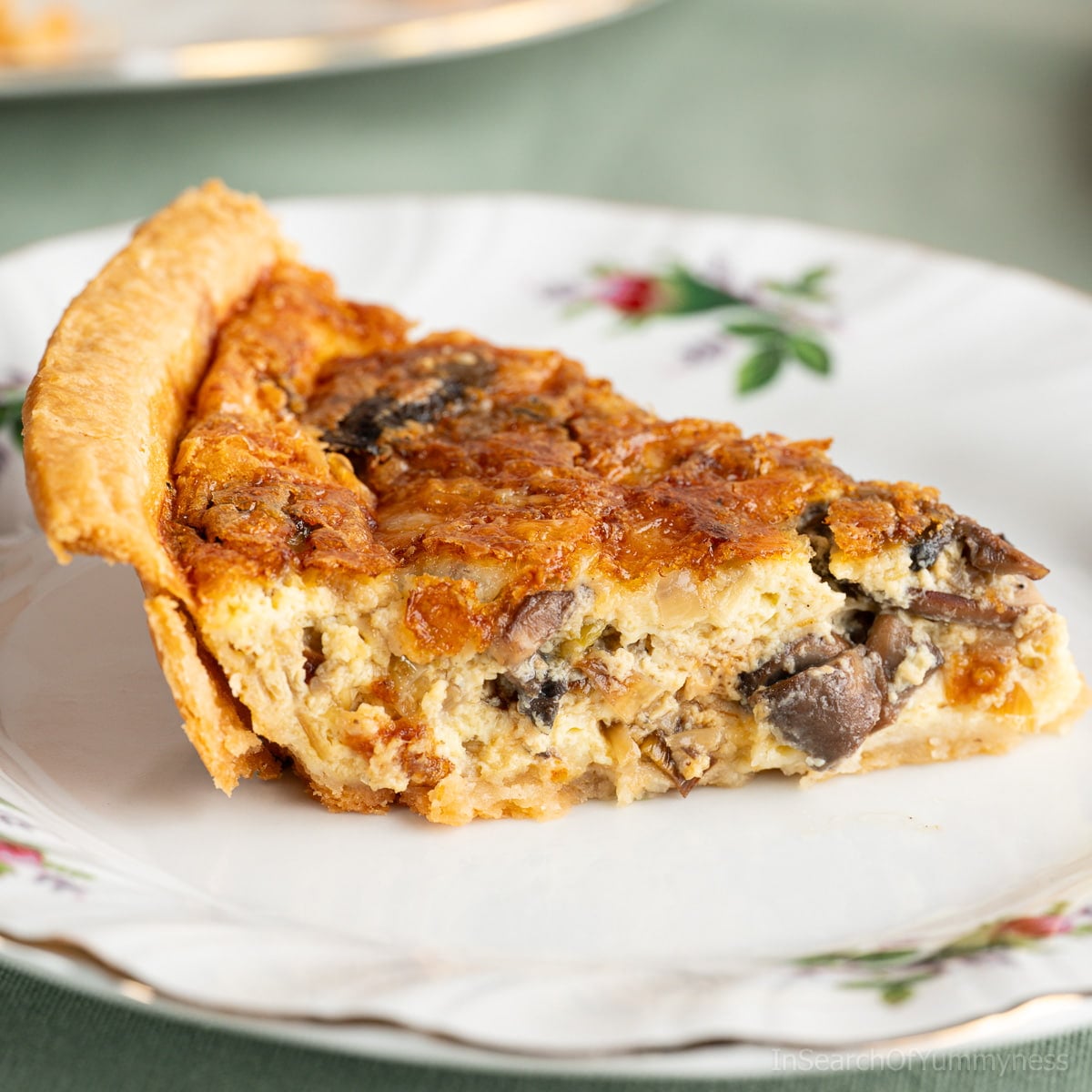 A slice of freshly baked leek and mushroom quiche with a cheddar cheese topping, sitting on a fancy white plate on a green tablecloth.
