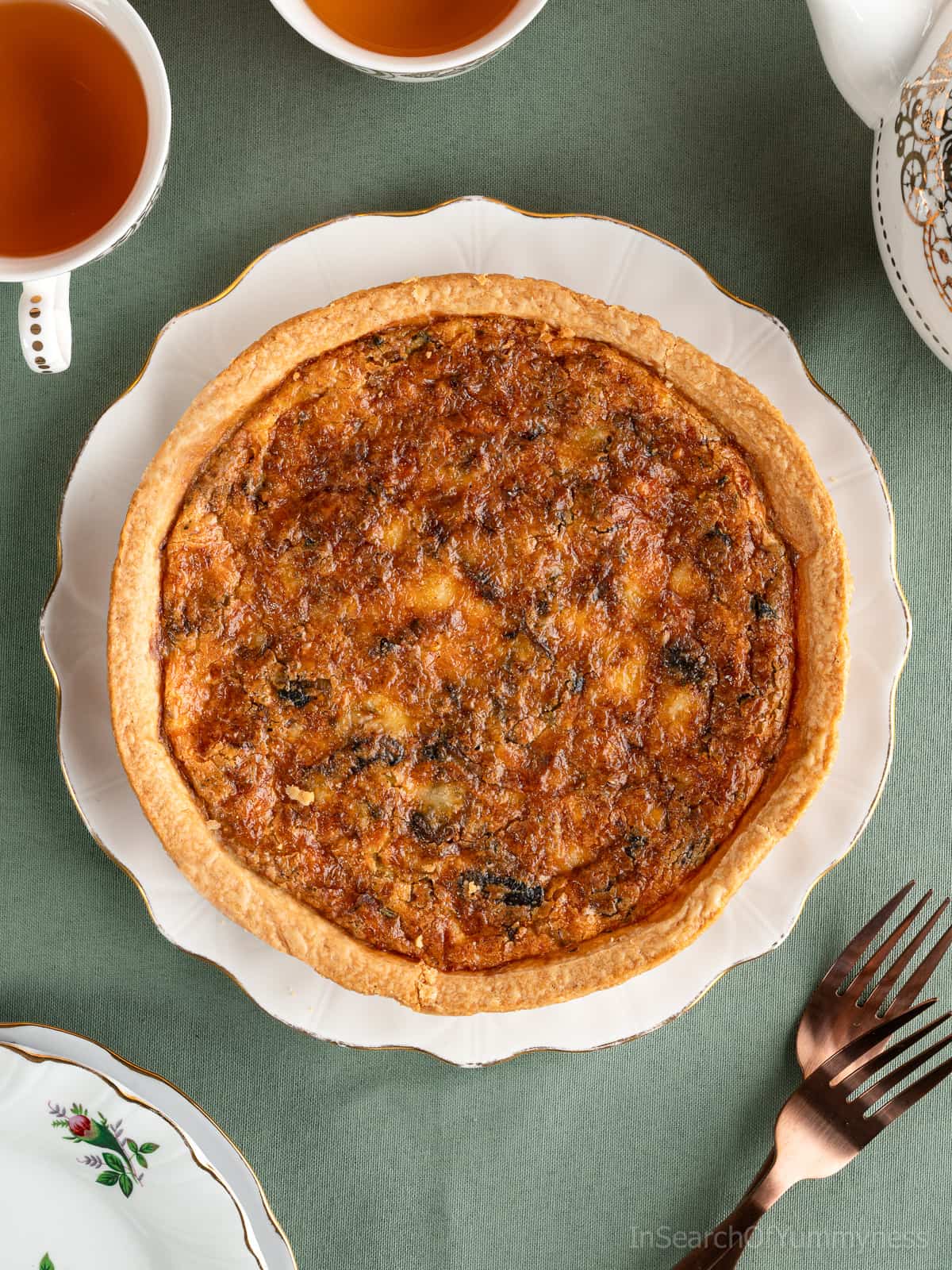 A baked leek and mushroom quiche with a crispy golden-brown cheddar topping, on a fancy white plate next to two teacups, a teapot, two forks and two saucers.