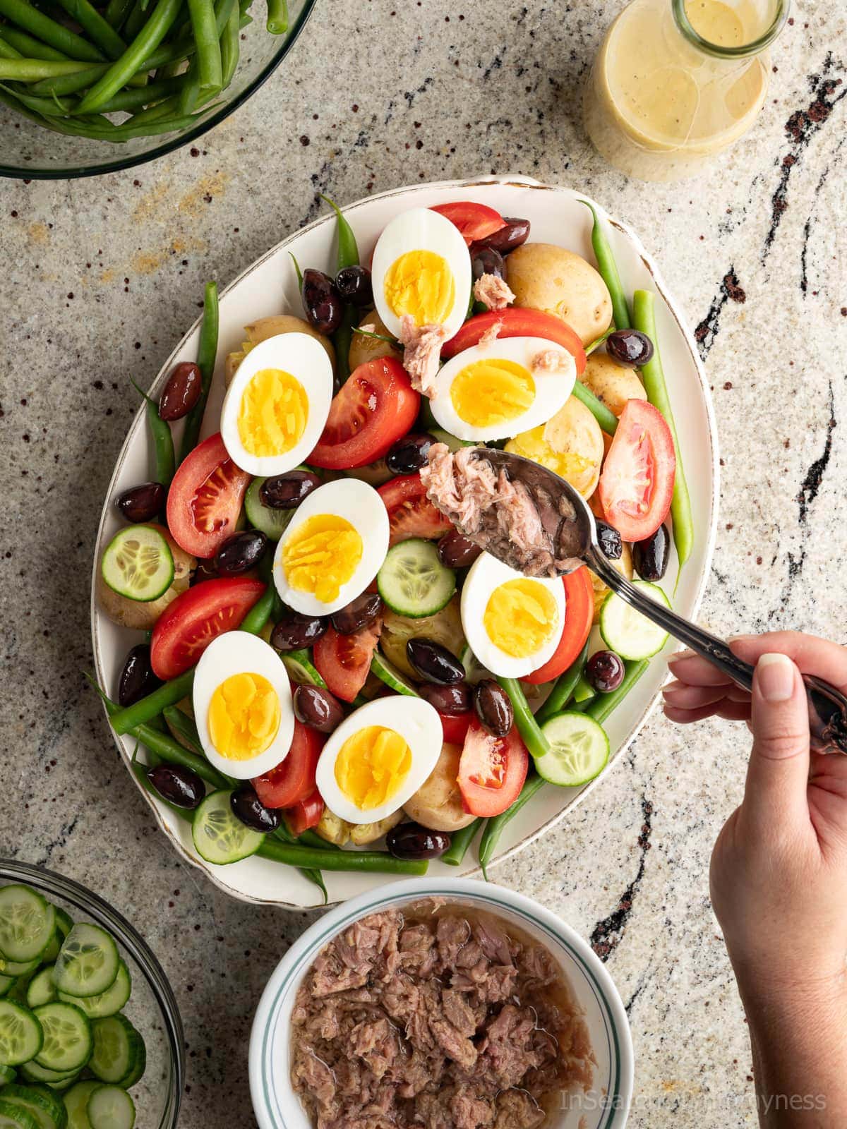 Someone using a spoon to add flaked tuna to a large oval platter with potatoes, cucumbers, olives, green beans, tomatoes and halved boiled eggs, sitting next to a glass bottle of lemony dressing and a ceramic bowl of canned tuna.