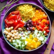 A bowl of couscous salad with chickpeas, tomato wedges, and diced vegetables next to a wooden spoon on a purple napkin.