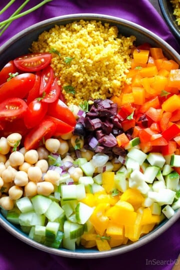 A bowl of couscous salad with chickpeas, tomato wedges, and diced vegetables next to a wooden spoon on a purple napkin.