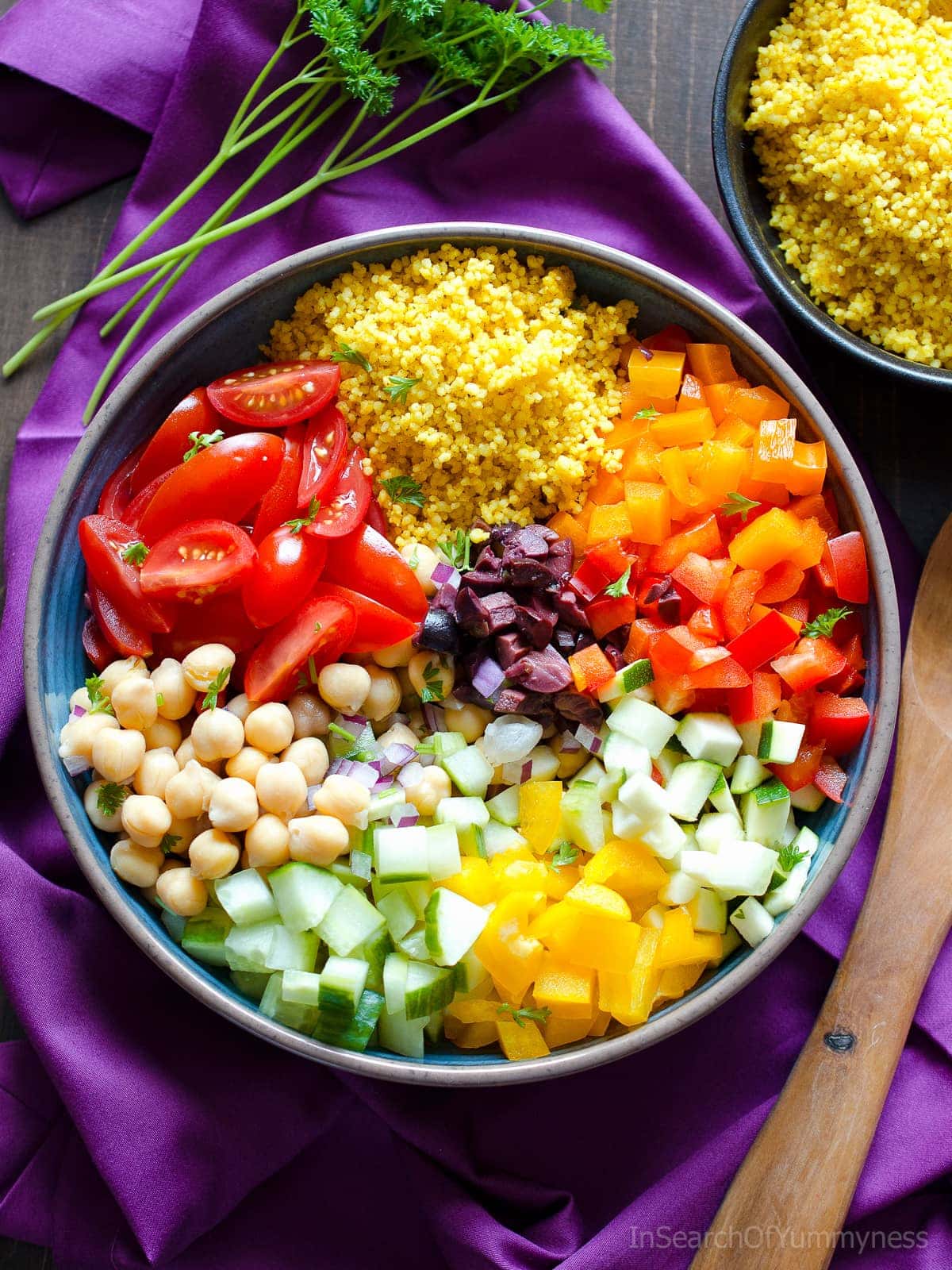 A bowl of couscous salad with chickpeas, tomato wedges, and diced vegetables next to a wooden spoon on a purple napkin. There is another bowl of couscous on the top right.