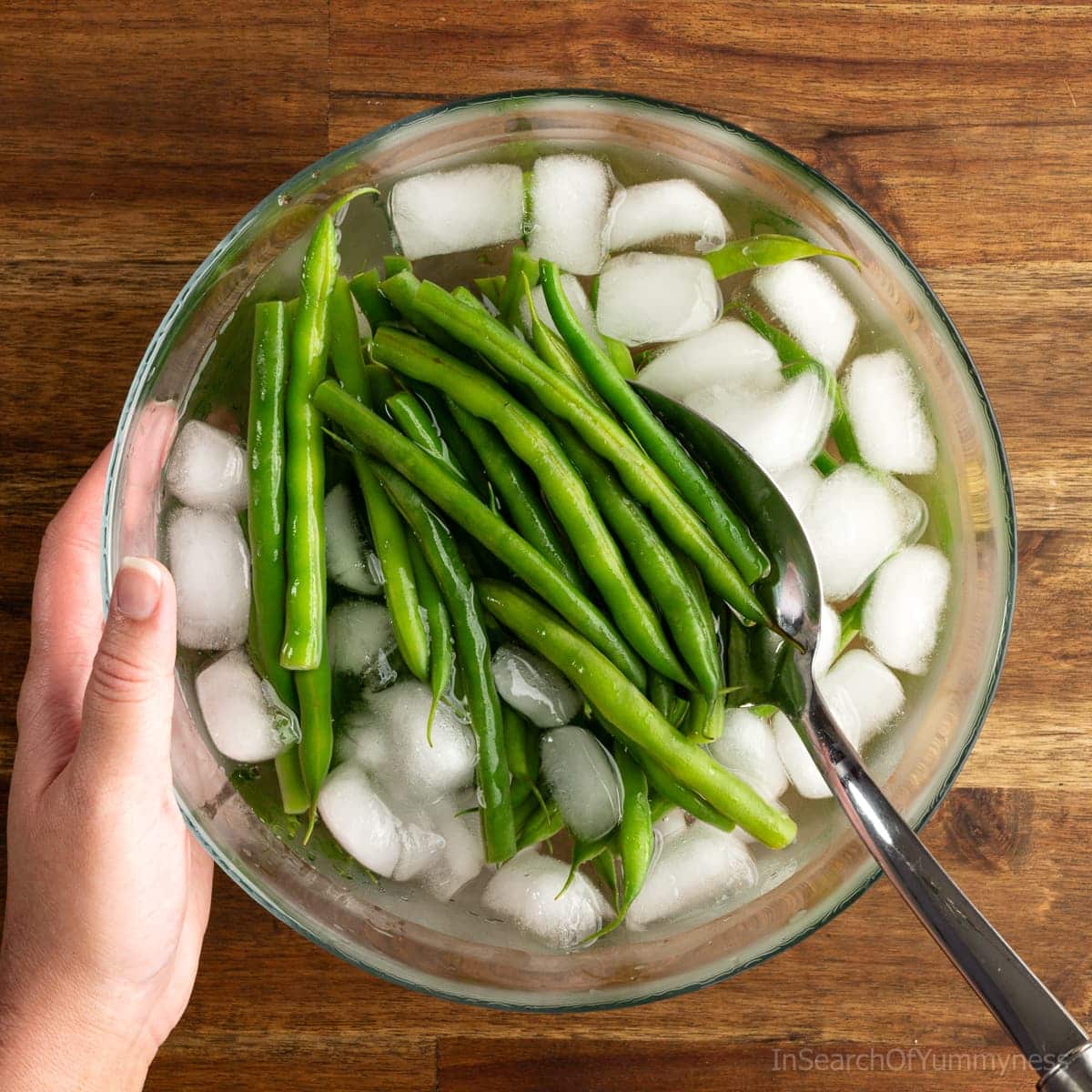 A slotted spoon lowering cooked green beans into a bowl of ice water to cool down.