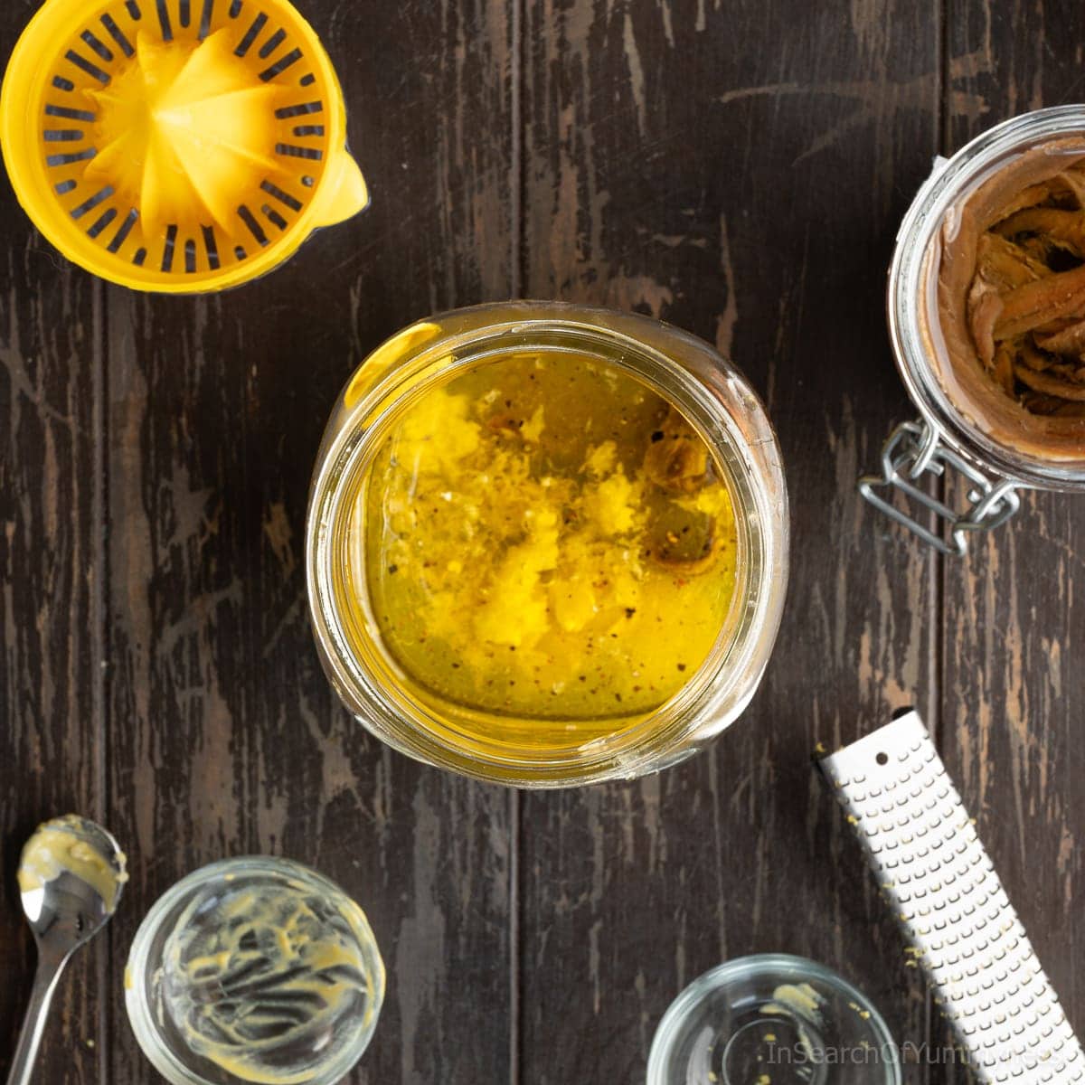 The ingredients for a lemony nicoise salad dressing in a wide-mouth mason jar, next to a jar of anchovies and empty glass bowls.