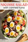 A large oval platter of Tuna Nicoise Salad with potatoes, sitting next to two amber drinking glass, a glass bottle of lemony dressing and wooden salad servers. Text overlay says "Nicoise Salad with canned tuna."