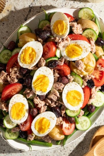 A large oval platter of Tuna Nicoise Salad with potatoes, sitting next to an amber drinking glass and wooden salad servers.
