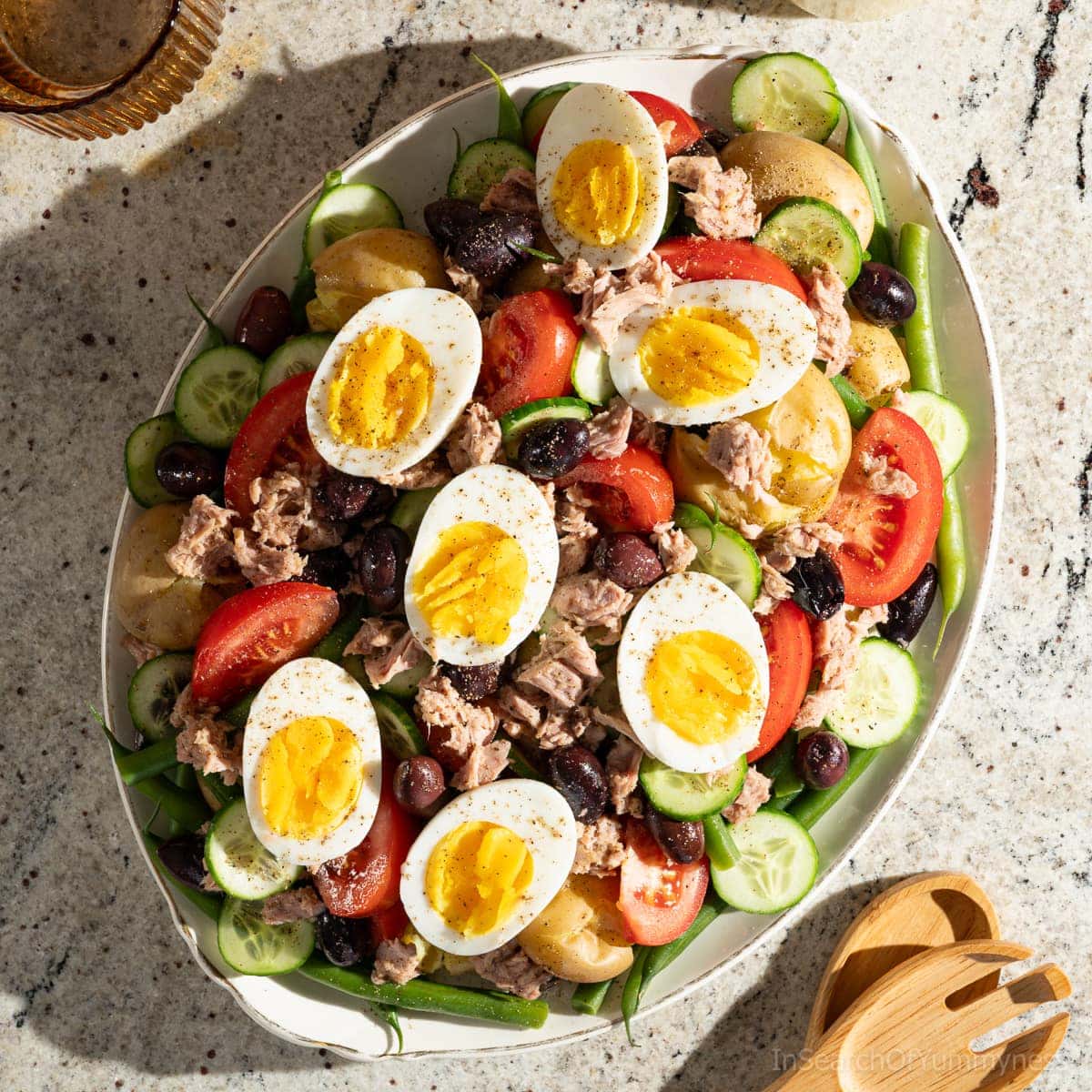 A large oval platter of Tuna Nicoise Salad with potatoes, sitting next to an amber drinking glass and wooden salad servers.