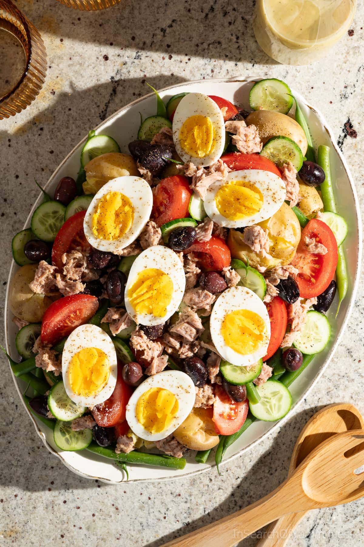 A large oval platter of Tuna Nicoise Salad with potatoes, sitting next to two amber drinking glasses, a glass bottle of lemony dressing and wooden salad servers.